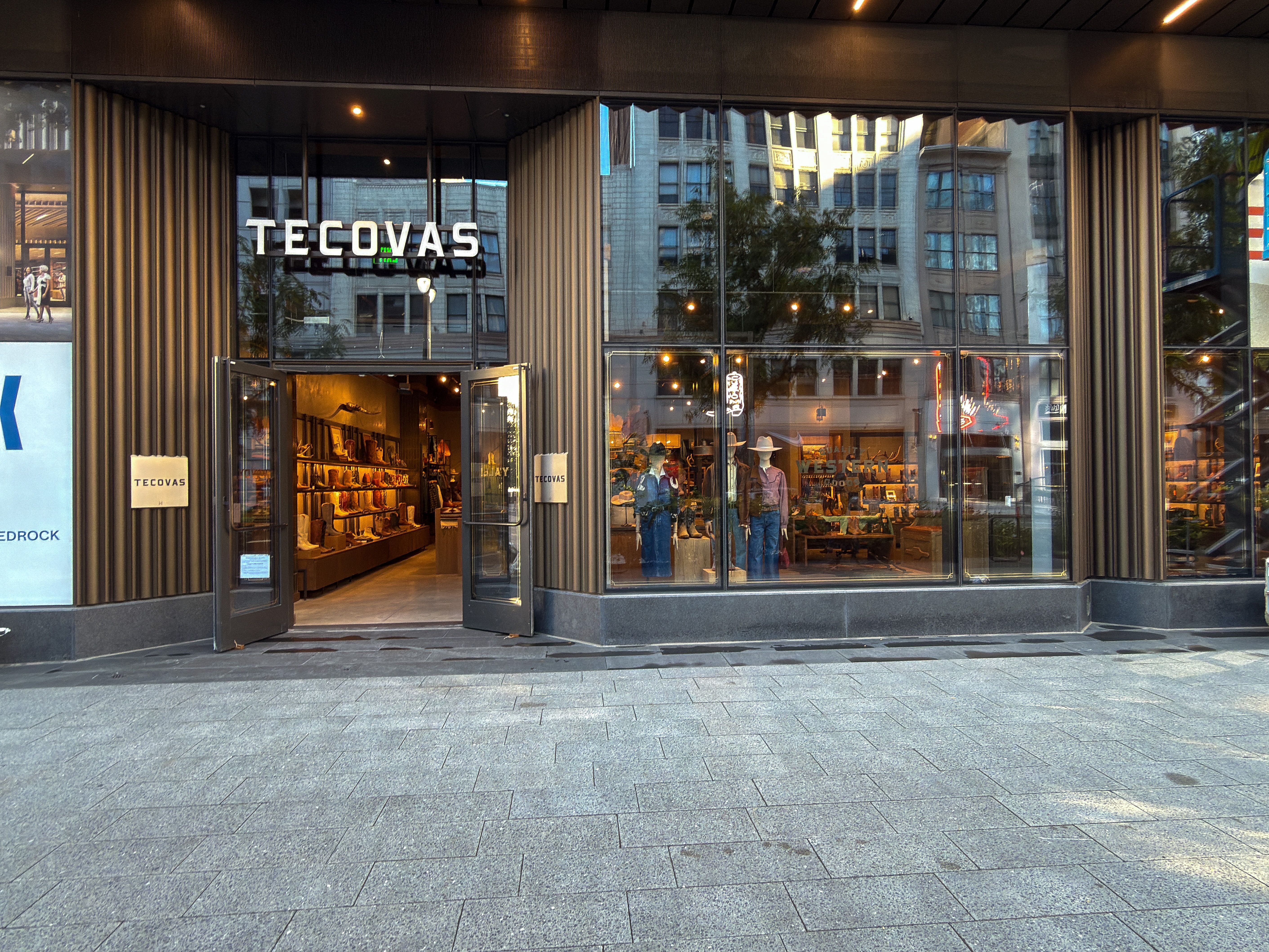 Street view of a Tecovas store with open glass doors, showcasing boots and accessories inside; two people are visible browsing the merchandise.