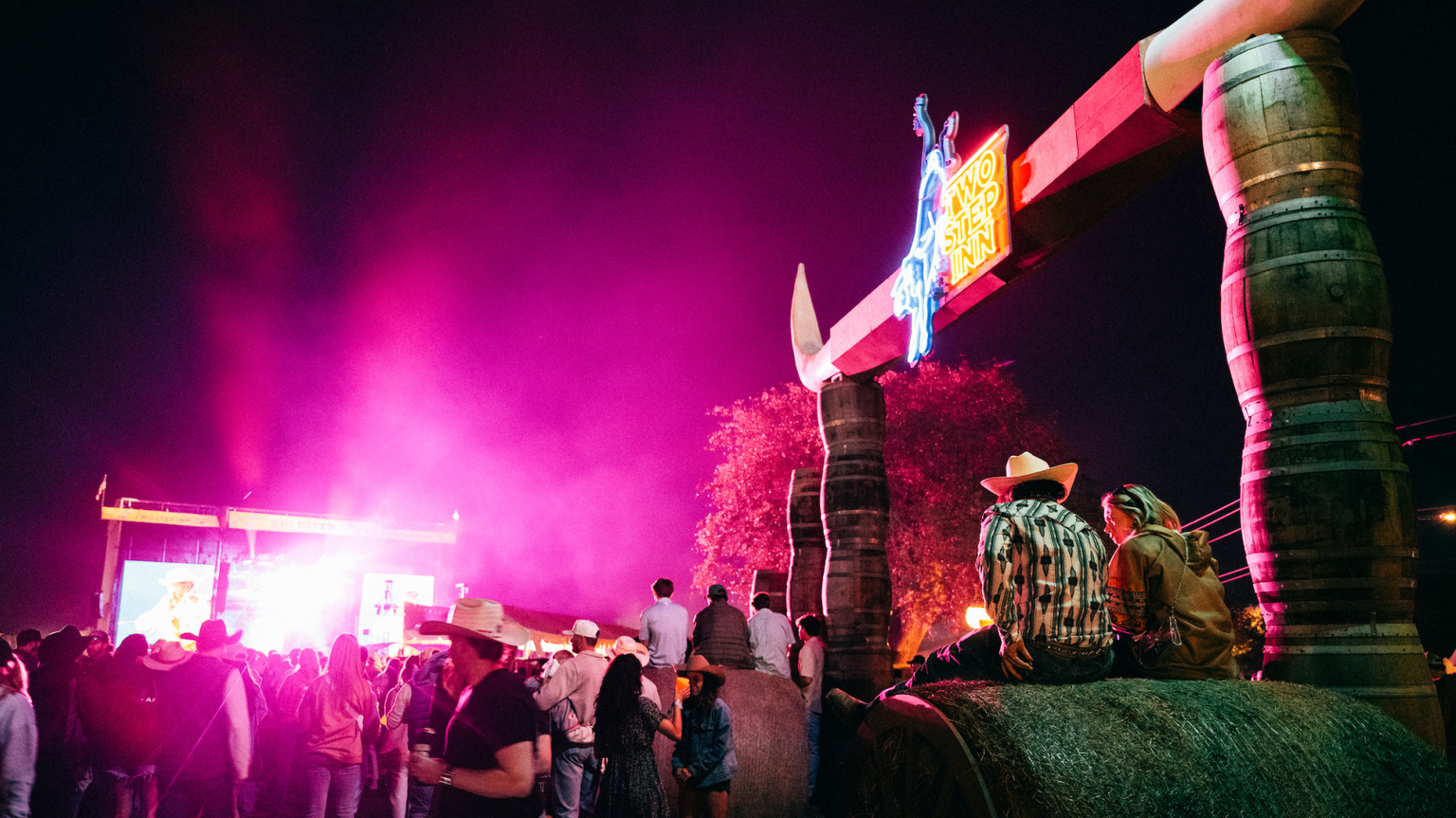 People gathered at an outdoor night event, with neon lights and a sign featuring large tusks. Two individuals in cowboy hats sit on a structure, while a stage is visible in the background.