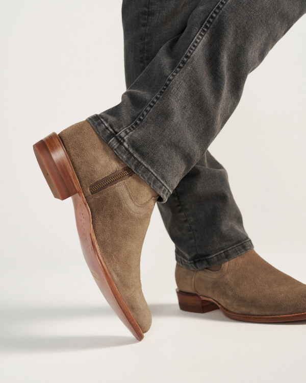 A person wearing gray jeans and The Dean Suede ankle boots in Granite, featuring light brown suede and wooden soles, stands against a plain background.