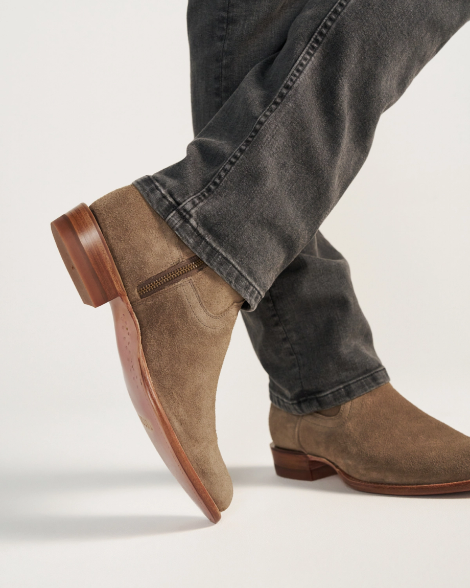 A person wearing gray jeans and The Dean Suede ankle boots in Granite, featuring light brown suede and wooden soles, stands against a plain background.