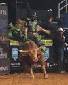 A bull rider wearing a helmet and protective vest rides a bucking bull in an indoor rodeo arena, with dirt flying and spectators in the background.