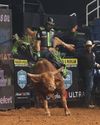 A bull rider wearing a helmet and protective vest rides a bucking bull in an indoor rodeo arena, with dirt flying and spectators in the background.