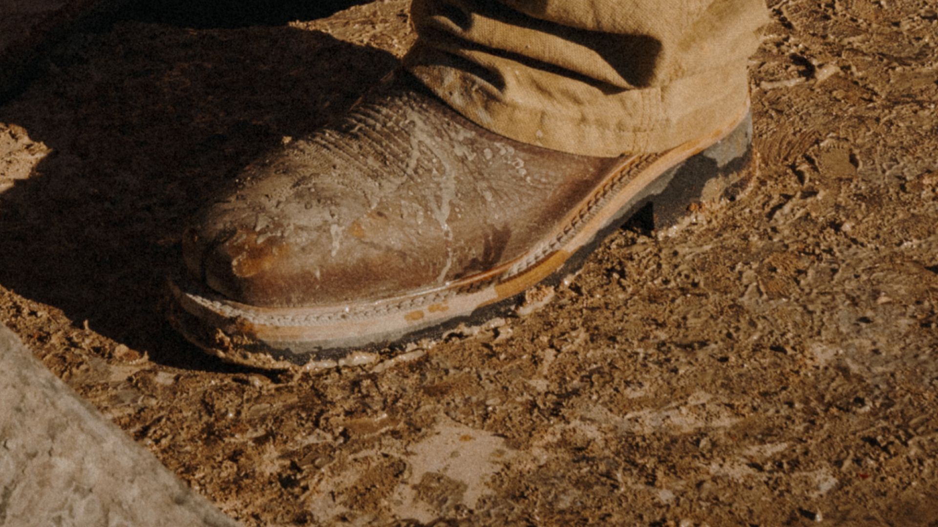 A close-up of a brown leather work boot and tan pants, both covered in dried mud, standing on a muddy, textured ground.