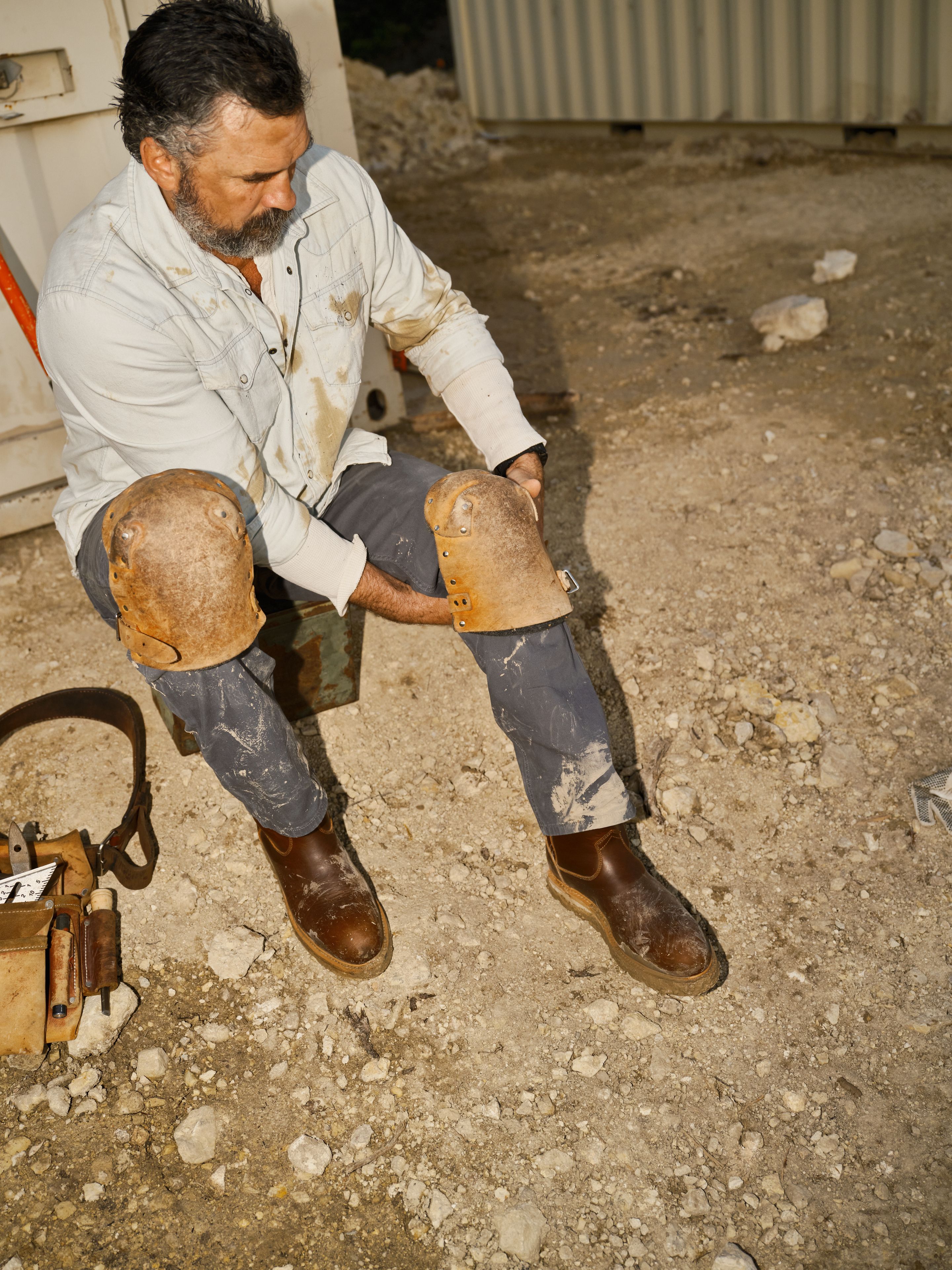 A man sitting on the ground at a construction site puts on heavy-duty knee pads over his jeans, with a tool belt beside him.