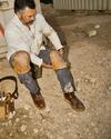 A man sitting on the ground at a construction site puts on heavy-duty knee pads over his jeans, with a tool belt beside him.