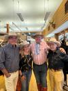 Four people wearing cowboy hats and casual attire are standing together in a western-themed store. Mounted deer heads and shelves with various items are visible in the background.