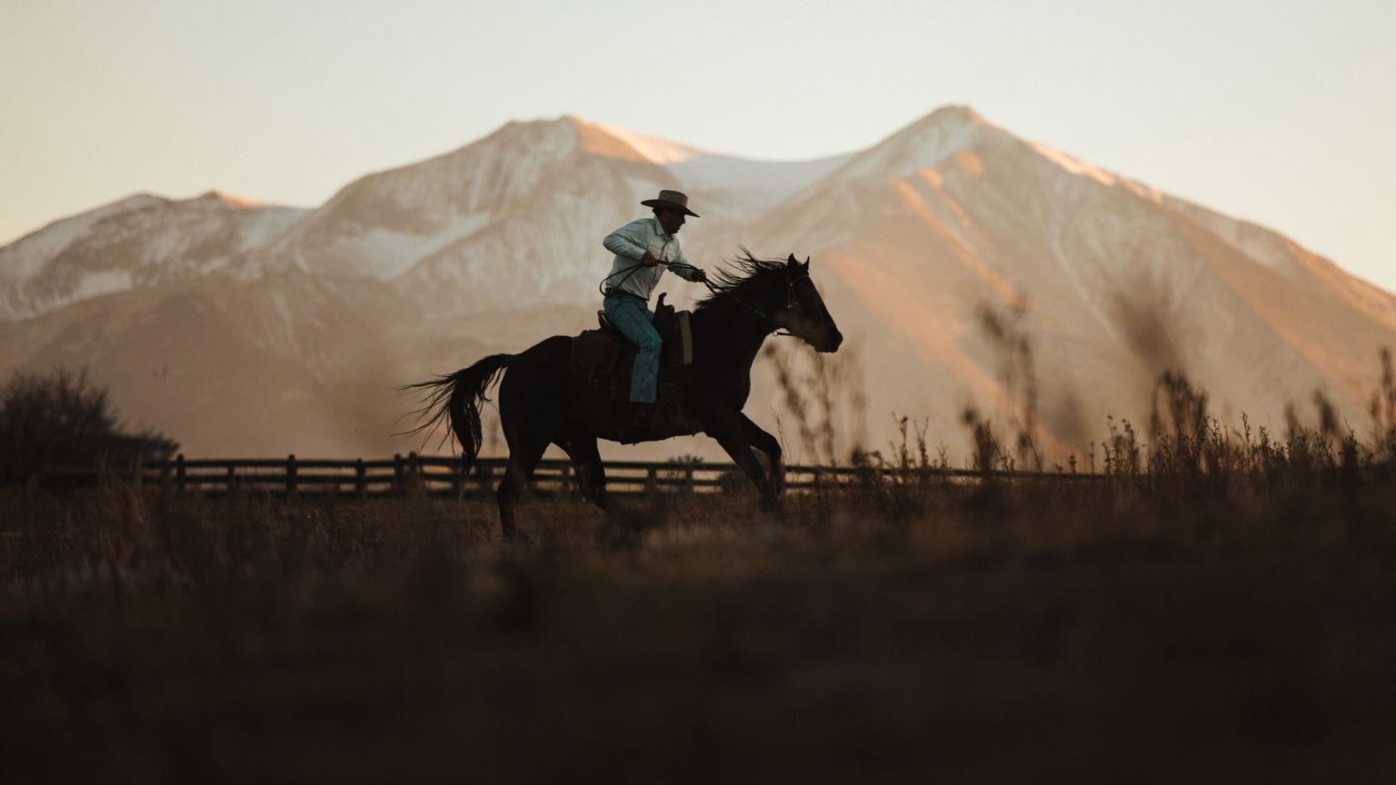A person wearing a cowboy hat rides a horse at a gallop in front of snow-capped mountains during sunset.