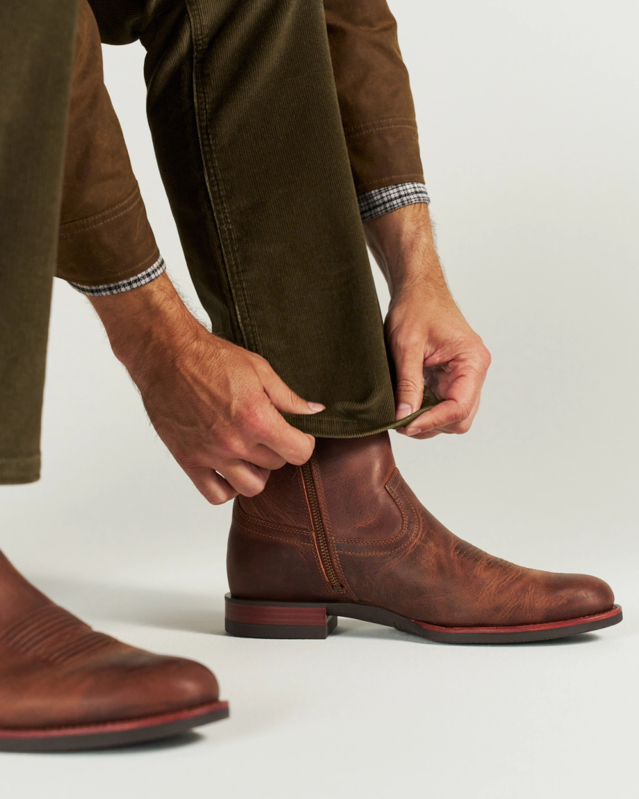 The Wayne in Maplewood Cowhide. A person wearing brown pants and plaid shirt cuffs tucks a pant leg into a Maplewood Cowhide brown leather boot with a red sole.