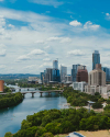 A cityscape view featuring a river with bridges and paddle boats, tall modern buildings, and green trees under a partly cloudy sky.