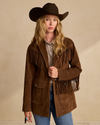 A woman in a cowboy hat and brown suede jacket with fringe details, standing against a beige background.