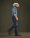 A man in a cowboy hat, denim shirt, jeans, and brown boots stands against a plain dark background, looking at the camera.