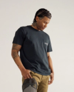 A man with braided hair and visible tattoos stands looking down, wearing a dark T-shirt with a chest pocket and khaki pants, holding a gray cap in one hand against a plain background.