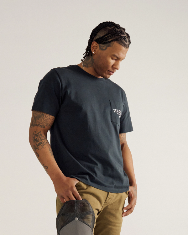 A man with braided hair and visible tattoos stands looking down, wearing a dark T-shirt with a chest pocket and khaki pants, holding a gray cap in one hand against a plain background.