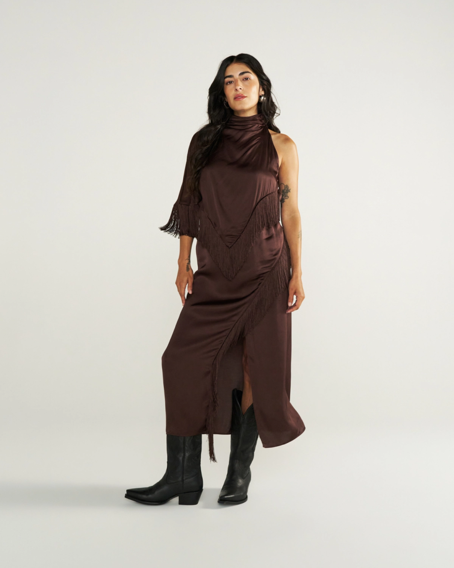 Woman in a sleeveless, dark brown fringed dress and black boots stands against a plain light background.