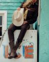 Person in Western-style clothing sits on an ice cooler, holding a cowboy hat, with turquoise-painted walls in the background.
