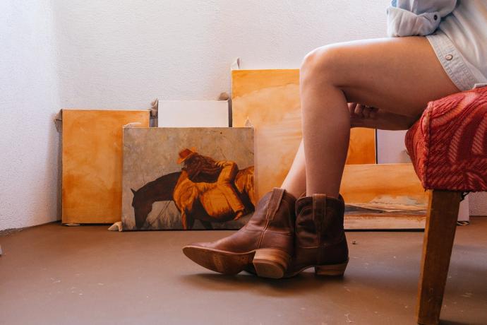 woman sitting on a chair wearing cowgirl booties with painted canvases in the background