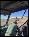 View from inside a vehicle showing a person leaning out while a cowboy on a horse rides across dry terrain, with a train passing in the background under a clear sky.