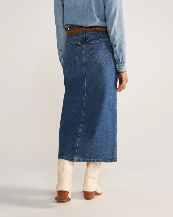 Person wearing a long denim skirt, brown belt, light blue denim shirt, and white cowboy boots, standing with their back to the camera.
