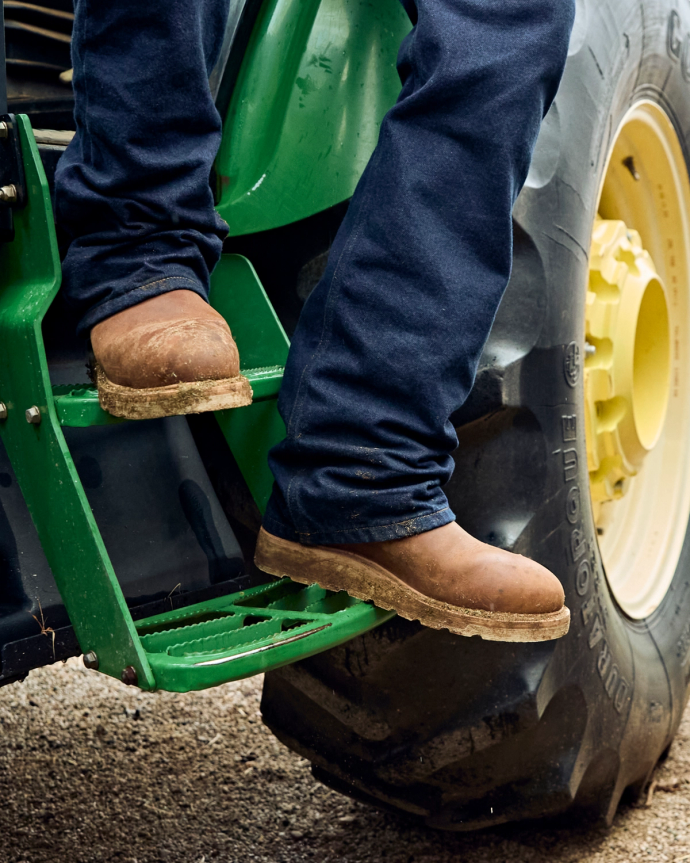 Person wearing brown boots and dark jeans steps onto a green tractor, with a large rear tire visible.