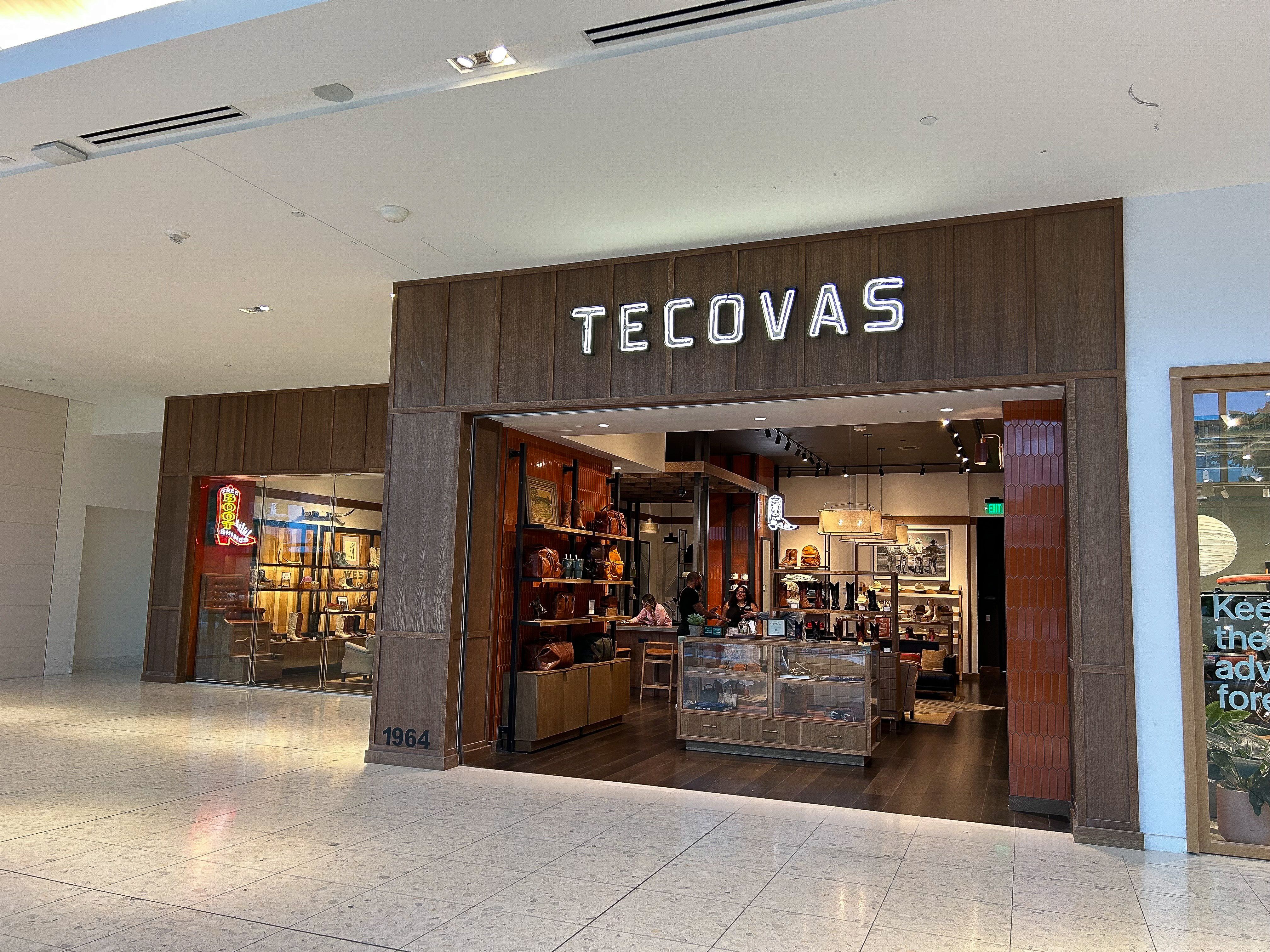 Storefront of Tecovas in a shopping mall, featuring leather goods and boots displayed inside, with glass walls and large illuminated signage above the entrance.