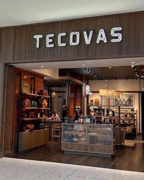 Storefront of Tecovas in a shopping mall, featuring leather goods and boots displayed inside, with glass walls and large illuminated signage above the entrance.