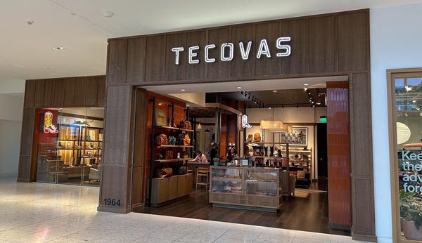 Storefront of Tecovas in a shopping mall, featuring leather goods and boots displayed inside, with glass walls and large illuminated signage above the entrance.