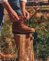 Person tying the laces of a brown boot while resting their foot on a tree stump in an outdoor setting.