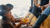 A person in a cowboy hat polishes white boots worn by another person sitting and holding a drink. Another person sitting nearby wears black boots.