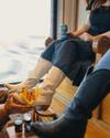 A person in a cowboy hat polishes white boots worn by another person sitting and holding a drink. Another person sitting nearby wears black boots.