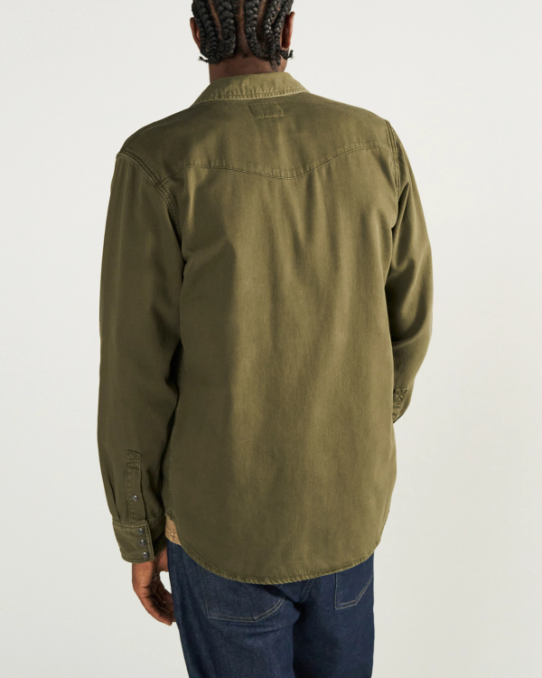 A person with braided hair is shown from behind, wearing an olive green long-sleeve shirt and dark blue jeans against a plain light background.