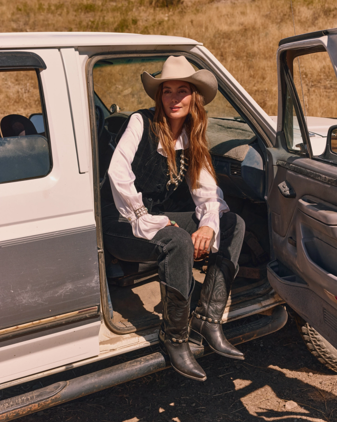 A woman in Western attire, including a cowboy hat, sits on the edge of a pickup truck's open door in an outdoor setting.