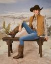 Woman in cowboy hat, puffer vest, button down shirt, jeans and cowgirl boots sitting on a wooden bench.