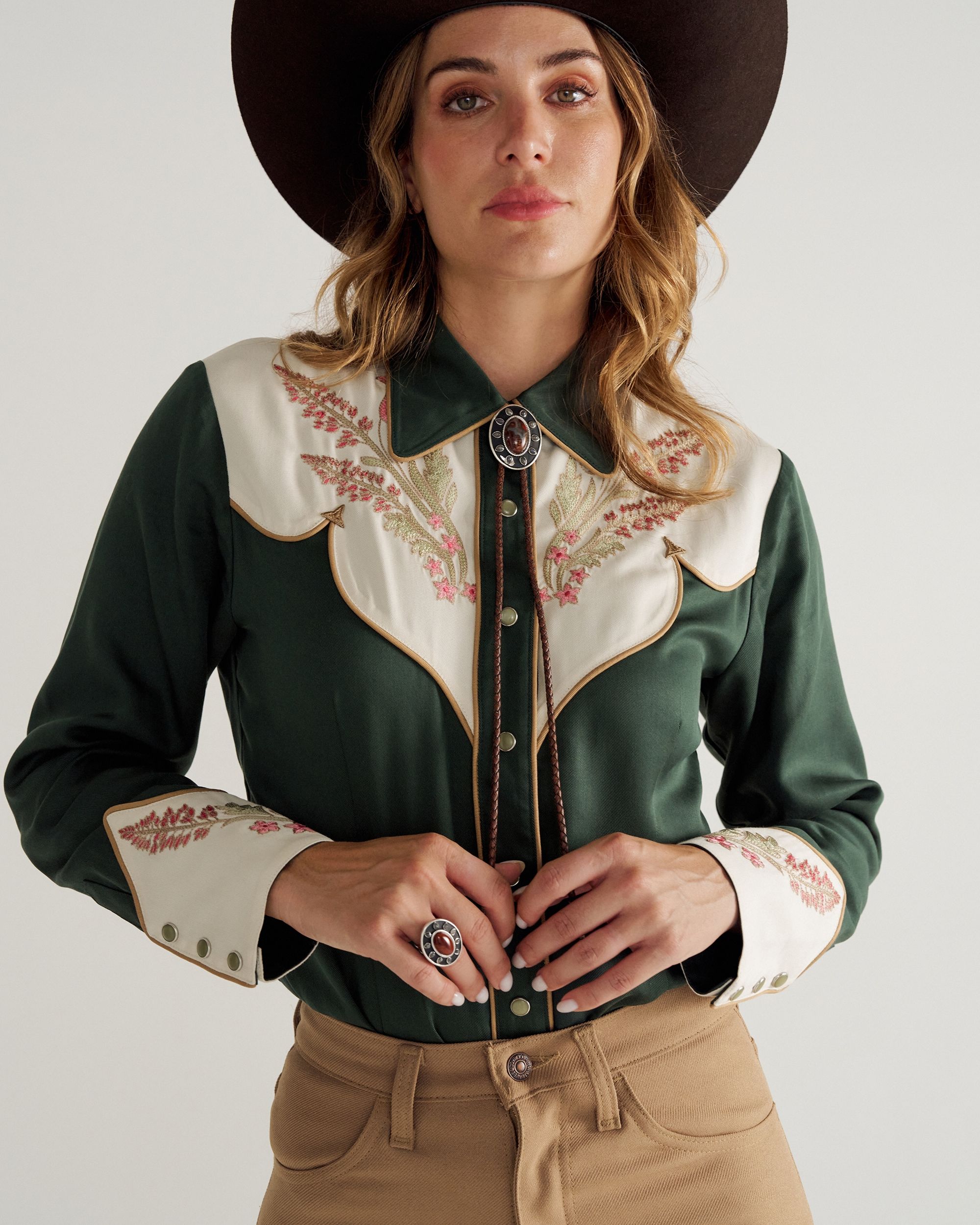 Woman wearing a dark green Western-style shirt with embroidered details, beige pants, and a brown cowboy hat, looking at the camera.