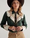 Woman wearing a dark green Western-style shirt with embroidered details, beige pants, and a brown cowboy hat, looking at the camera.