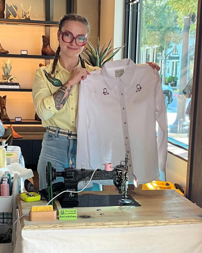 A person with braided hair and glasses stands behind a sewing machine, holding up a white shirt with embroidered designs in a brightly lit store.