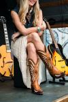 A woman sitting with her leg crossed, wearing a denim vest and decorative cowboy boots, in front of acoustic guitars.