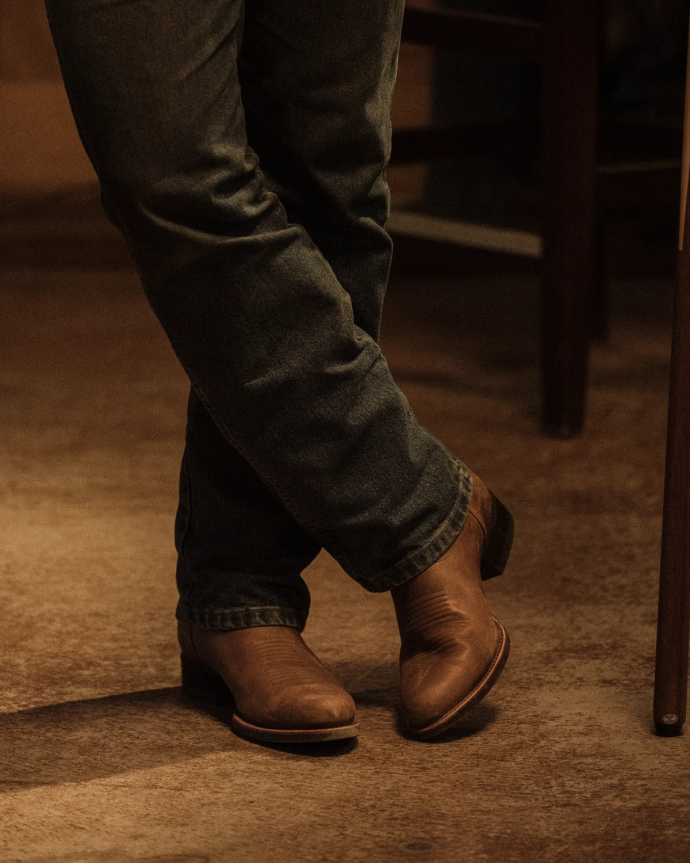 A person wearing brown leather boots and dark jeans stands with legs crossed on a wooden floor.