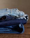 A neatly folded stack of three pairs of jeans in different denim shades placed on a wooden surface.