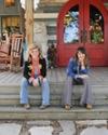 Two women sit on the steps of a wooden porch with red double doors behind them, flanked by decorative statues and rocking chairs.