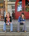 Two women sit on the steps of a wooden porch with red double doors behind them, flanked by decorative statues and rocking chairs.