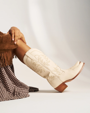 The Annie in Bone Cowhide. A person sits with legs crossed, wearing a brown fringed jacket, a checkered skirt, and The Annie Cowhide embroidered cowboy boots with wooden heels against a neutral background.