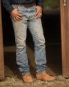 Person standing in a doorway wearing light blue jeans, a denim shirt, a brown belt, and tan shoes; hands resting on thighs, face not visible.