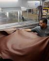 A man works with a large sheet of brown leather in an industrial setting, handling it near machinery.