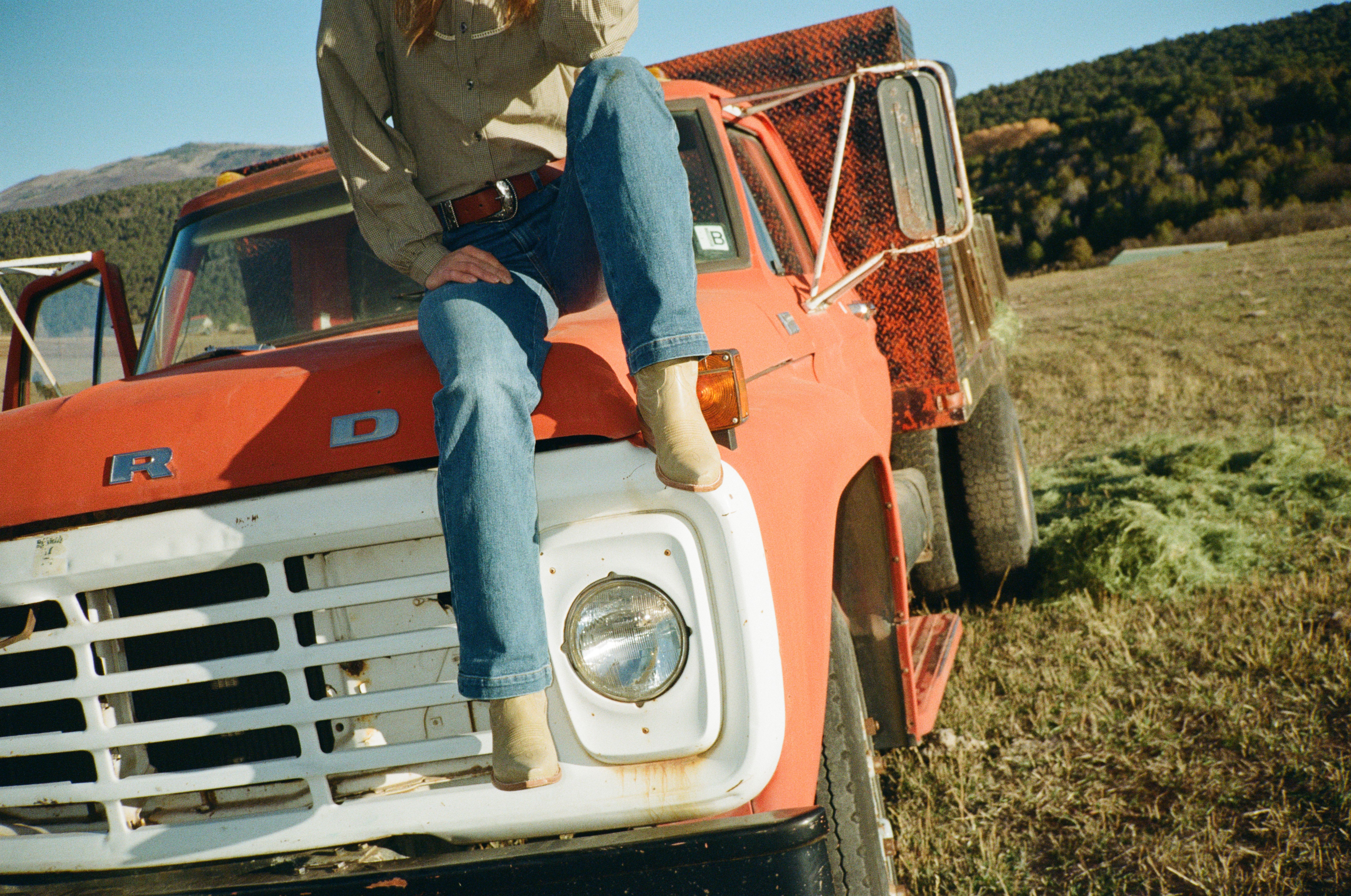 Person in jeans and boots sitting on the hood of an old red truck in a grassy field with mountains and trees in the background.