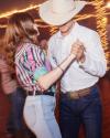 A man in a cowboy hat and white shirt dancing with a woman in a colorful striped shirt and jeans in a lively dance hall.