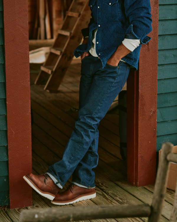 A man leaning against a doorway, wearing a quilted blue denim jacket, jeans, and The Knox Lace Up Cowhide boots in cognac.