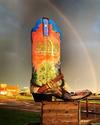 Large decorative cowboy boot with "Texas Boot Company" displayed on a wooden wagon with wheels, set on a grassy area with a rainbow in the background.