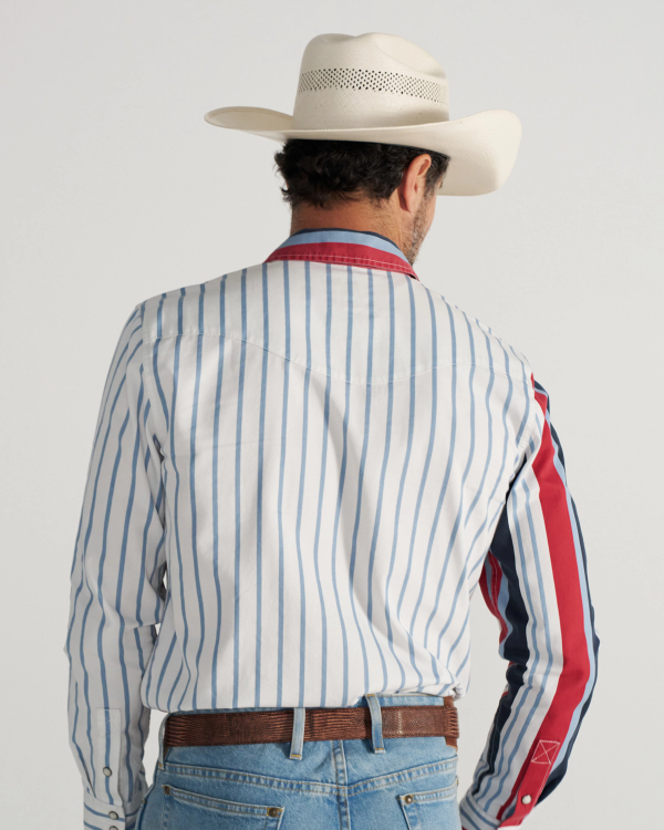 A man wearing a white cowboy hat, a striped long-sleeve shirt with red accents, blue jeans, and a brown belt stands facing away from the camera.