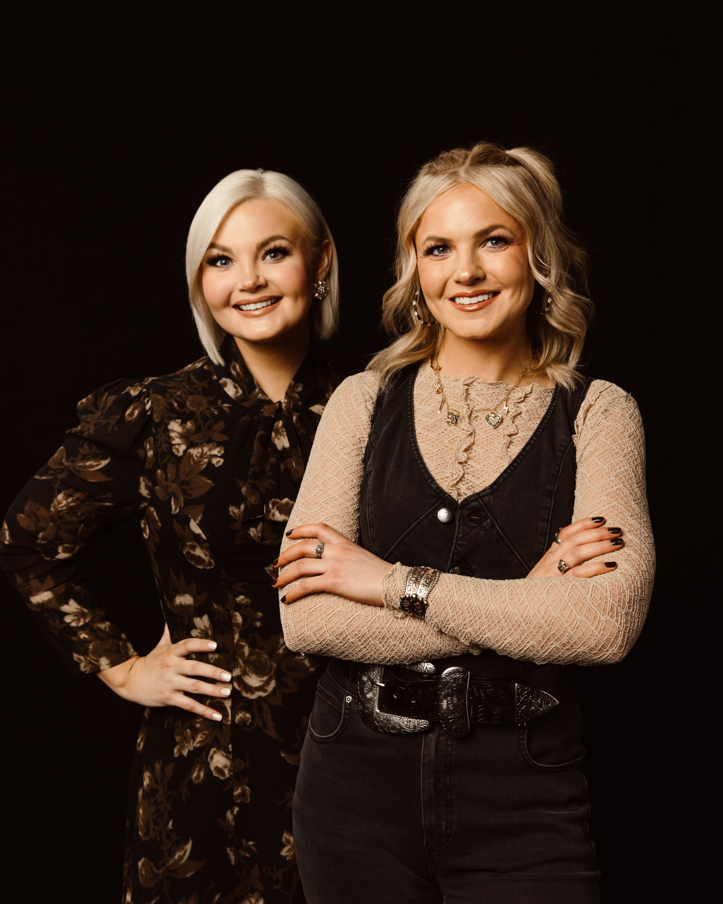 Two women stand in front of a black background. The woman on the left wears a floral dress, and the woman on the right wears a lace top with a black vest and pants. Both are smiling.
