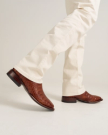 Person wearing light-colored pants and The William Caiman dark pecan crocodile-patterned leather shoes standing on a white surface.